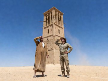 Observer shading their eyes while looking up at a tall mud-brick windcatcher tower against a clear sky